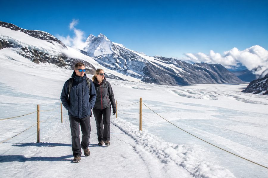 Dos excursionistas abrigados caminando por una meseta nevada de alta montaña junto a las cuerdas que marcan el paso